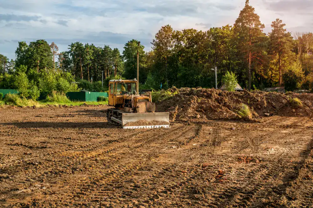 A yellow track bulldozer is operating on a large expanse of cleared, tilled brown dirt with tire tracks, leveling a pile of soil with its blade against a backdrop of dense green trees and a cloudy sky in Suffolk County, NY.